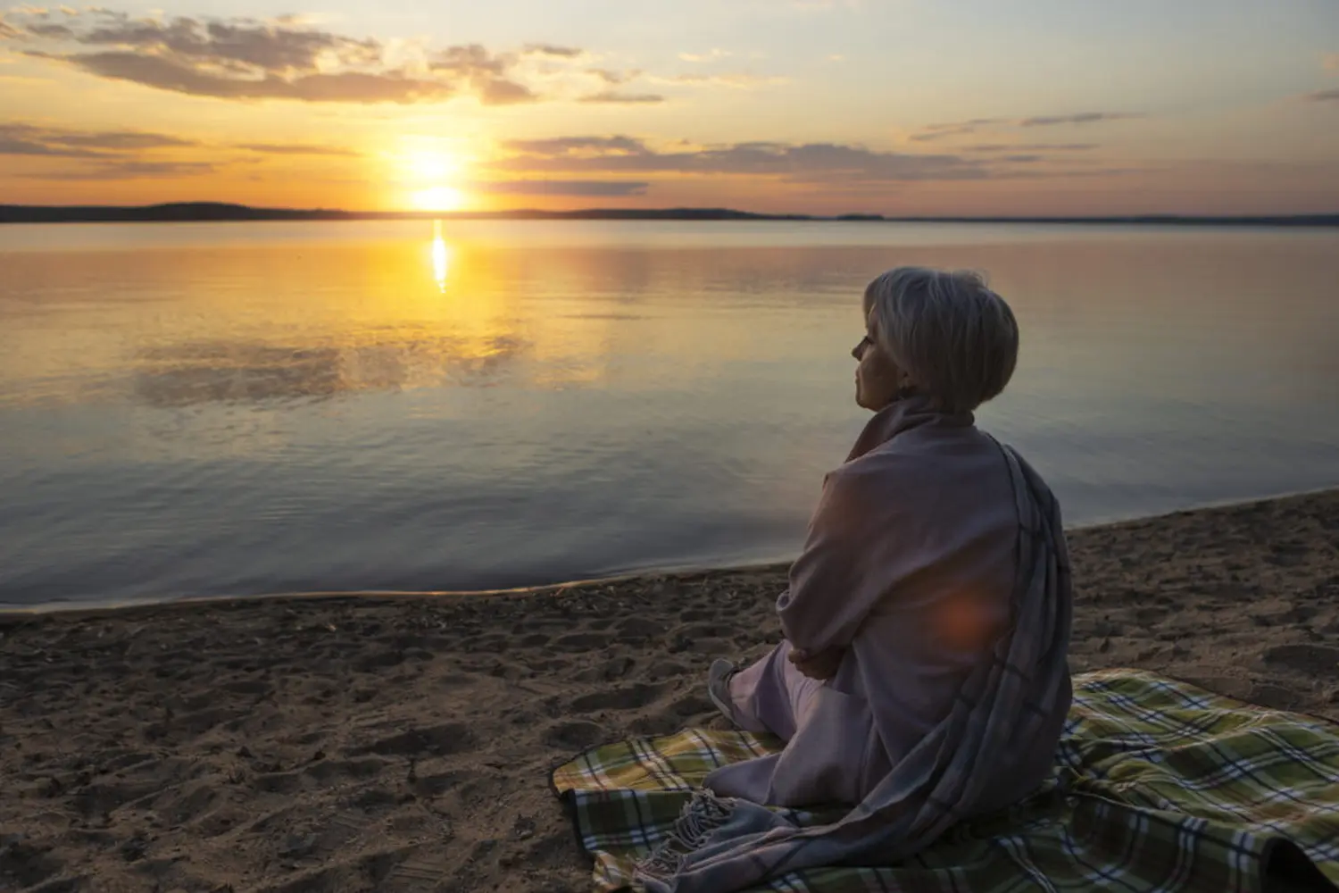 Eine Frau meditiert bei Sonnenuntergang am Strand. Sie sitzt auf einer Decke im Sand,